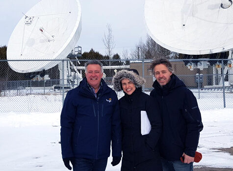 Three employees posing in front of two large satellite dishes connected to the Strigo satellite.